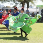 Three women in colorful traditional dresses dance outdoors on grass during a cultural festival, while spectators watch and a green tent is visible in the background.