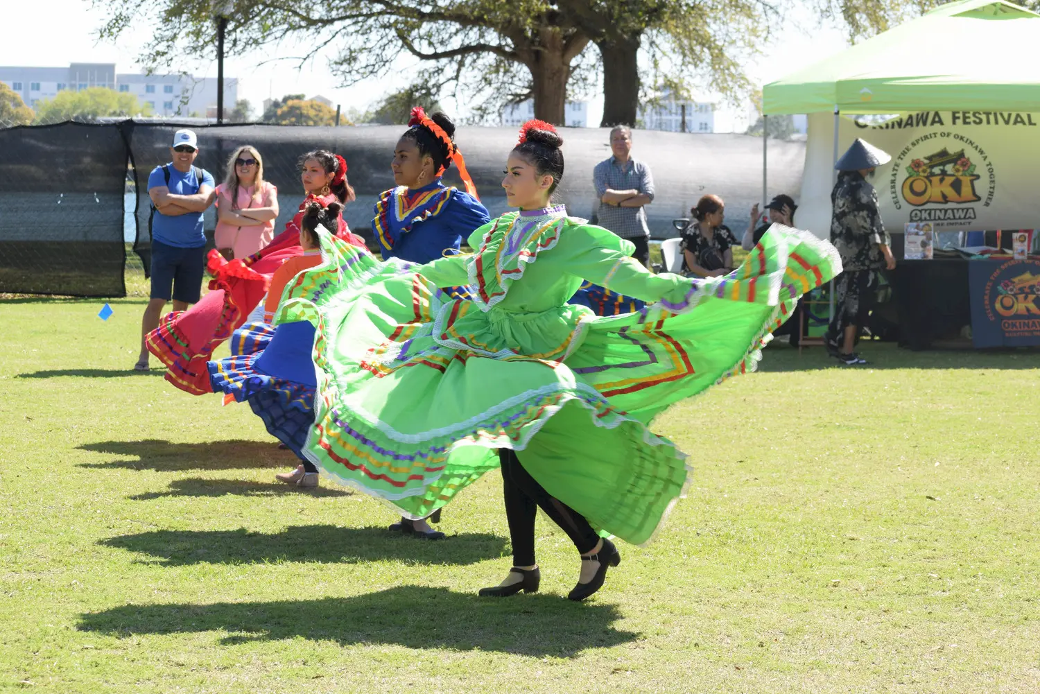 Three women in colorful traditional dresses dance outdoors on grass during a cultural festival, while spectators watch and a green tent is visible in the background.