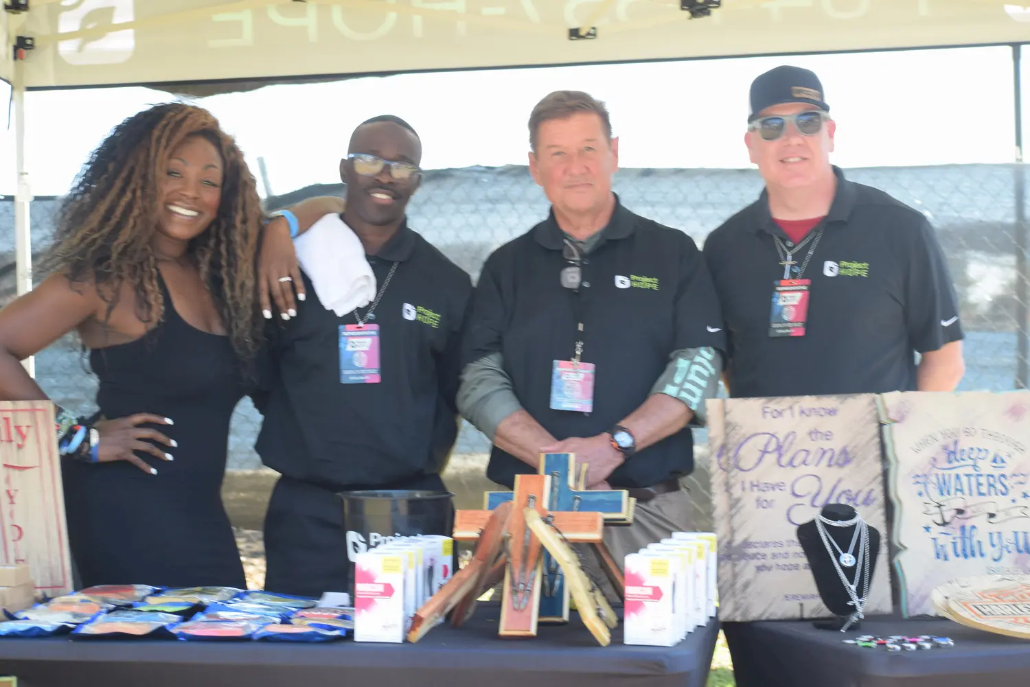 Four people stand smiling behind a booth table with various items such as snacks, wooden crosses, jewelry, and signs. They are outdoors, wearing badges and black shirts under a canopy.