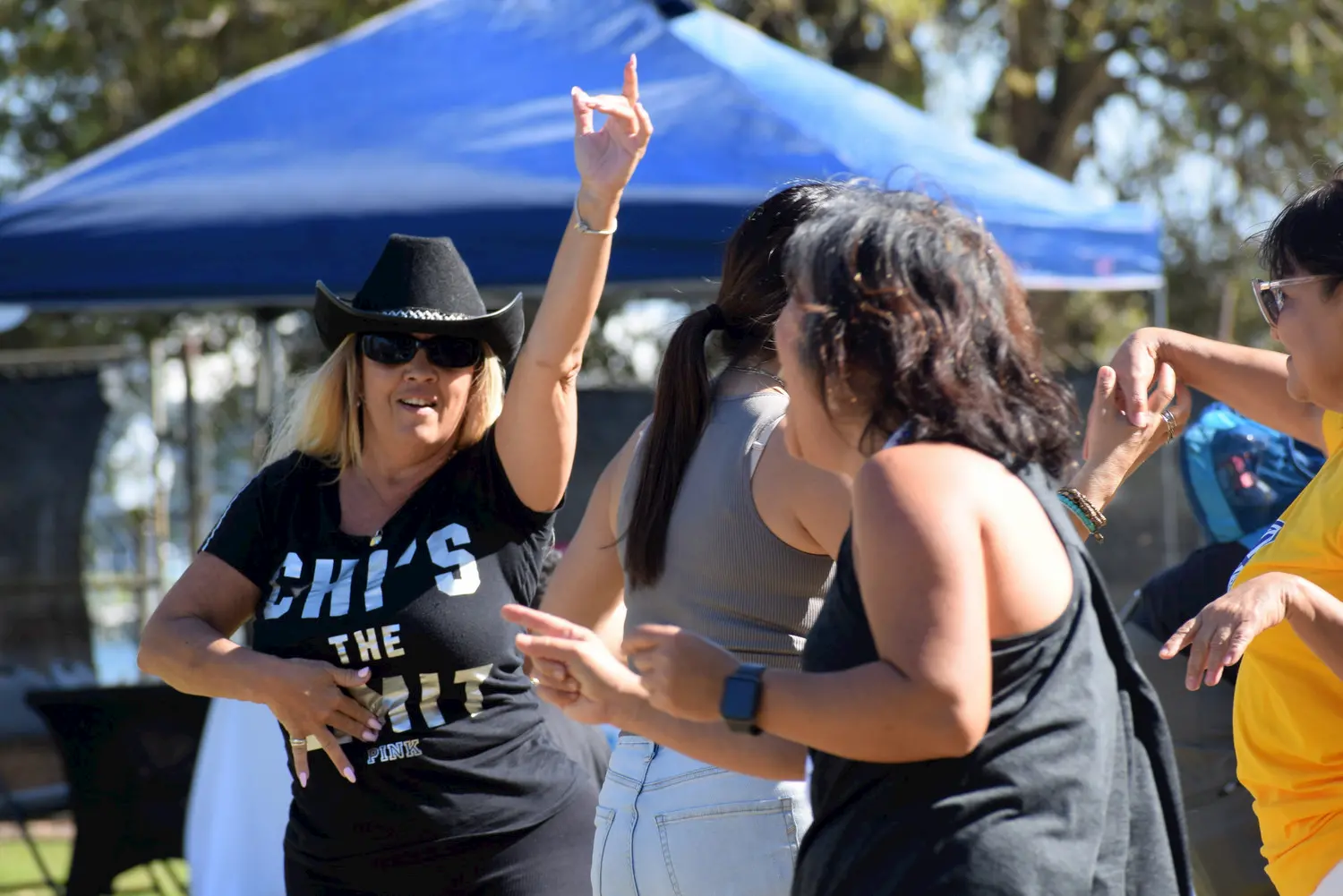 A group of women are dancing outdoors in the daytime, one wearing a black cowboy hat and sunglasses, with a blue canopy and trees in the background.