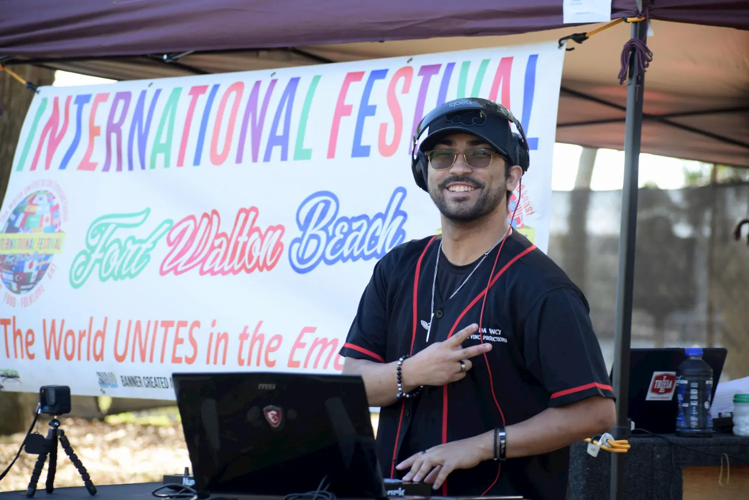 A smiling DJ wearing sunglasses and headphones stands at a booth with a laptop, flashing a peace sign. Behind him is a colorful "International Festival Fort Walton Beach" banner.