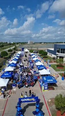 Aerial view of an outdoor event with white tents lined up on both sides of a street, groups of people gathered, and blue decorations featuring the Goya brand under a partly cloudy sky.