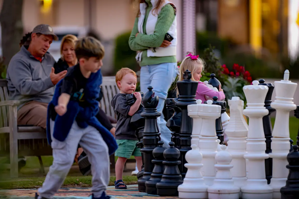 Children play with large black and white chess pieces outdoors, while adults sit on a bench in the background watching and using a phone. Flowers and greenery surround the scene.