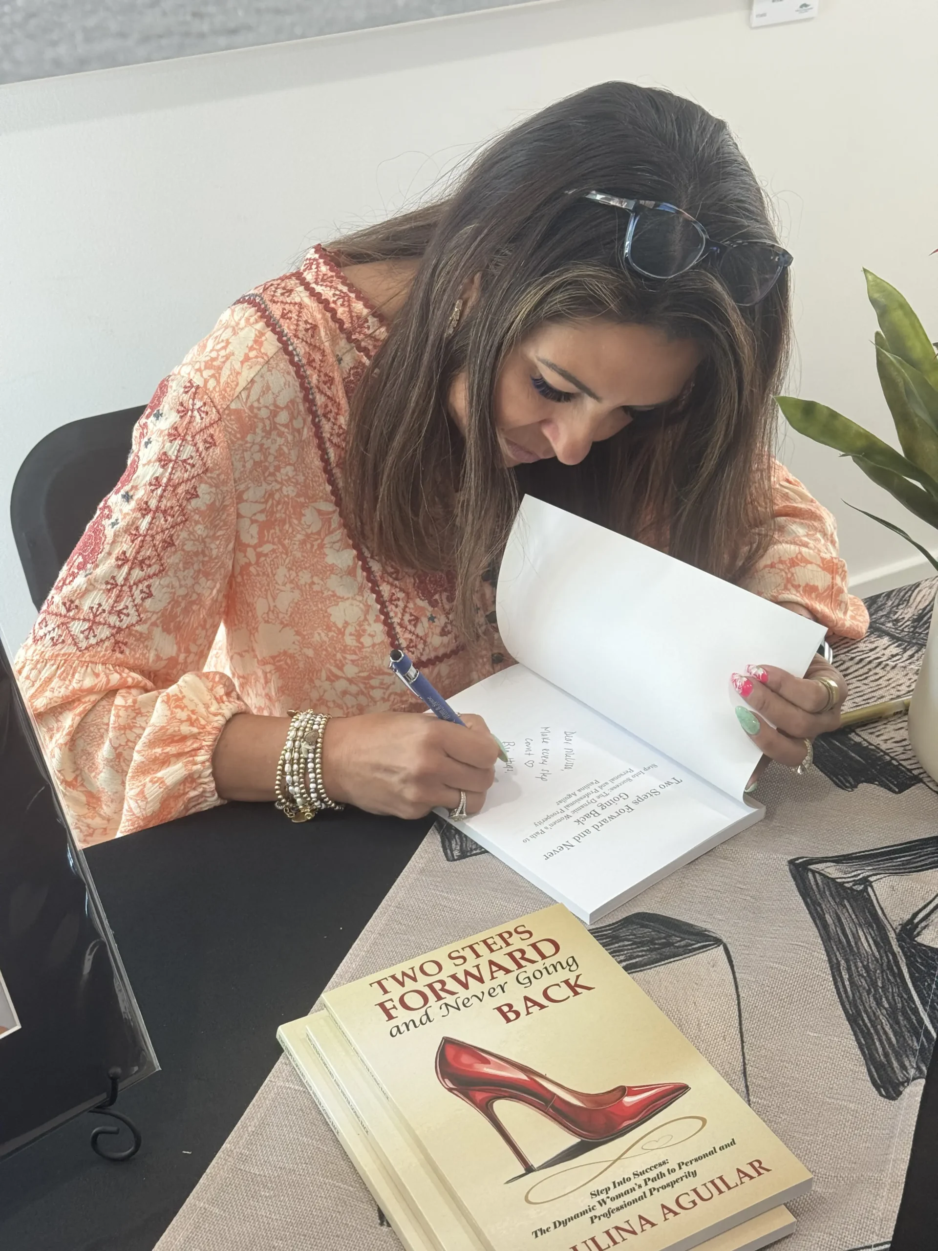 A woman with long brown hair and sunglasses on her head signs a book at a table. A copy of "Two Steps Forward and Never Going Back" is visible in the foreground next to a plant.