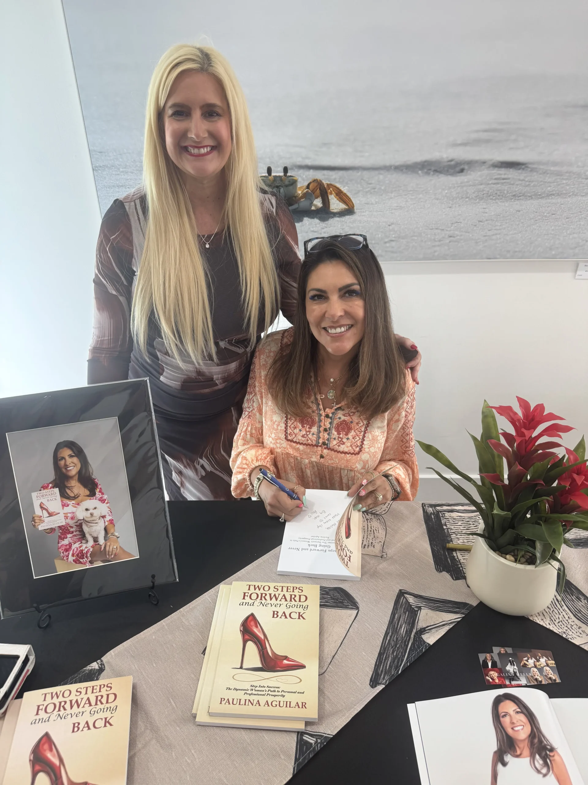Dos mujeres sonríen en un acto de firma de libros. Una está sentada firmando "Dos pasos adelante, un paso atrás", una historia sobre cómo mantenerse firme y establecer límites personales. Libros, una foto enmarcada y una planta de bromelia roja adornan la mesa ante ellas.