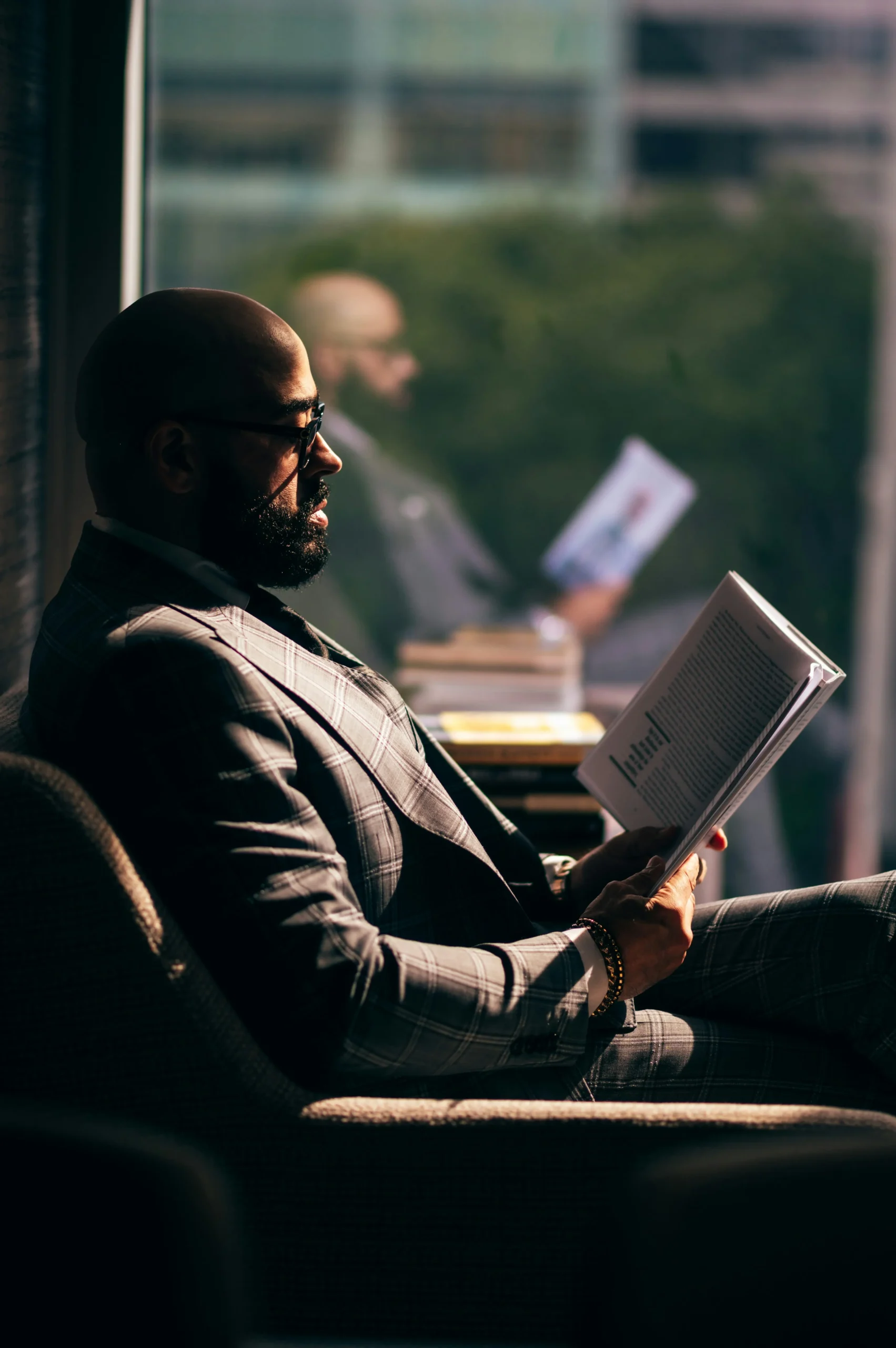 A man in a plaid suit sits in a chair by a window, reading a book. Sunlight streams in, casting a reflection of him holding the book in the glass behind him.