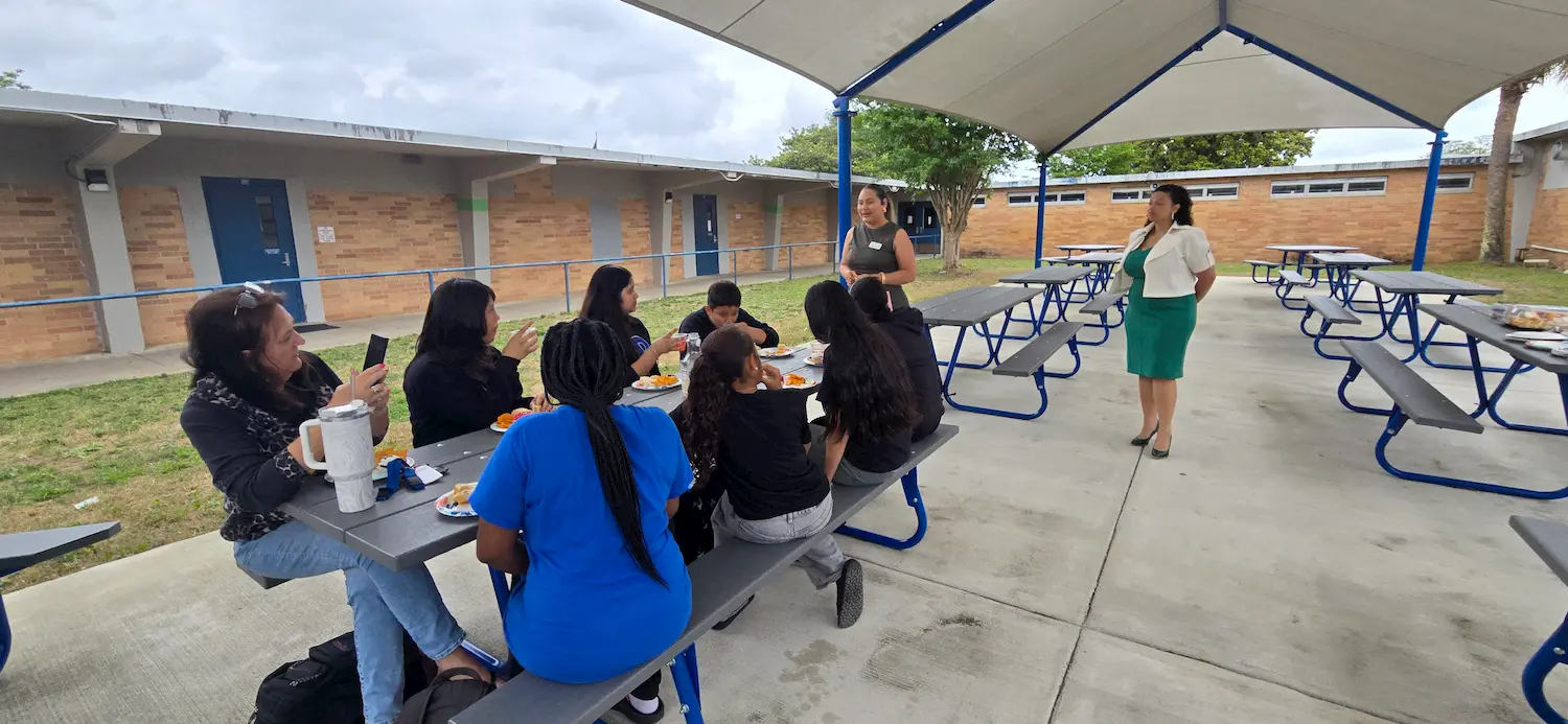 A group of people sit at picnic tables under a covered outdoor area, eating and talking, while two women stand nearby, one speaking to the group. The setting appears to be a school or campus courtyard.
