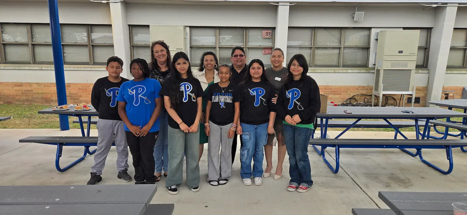 A group of students and adults pose together outdoors near blue picnic tables. Most of the students wear matching black shirts with a blue "R" logo. The adults stand behind the students, smiling at the camera.