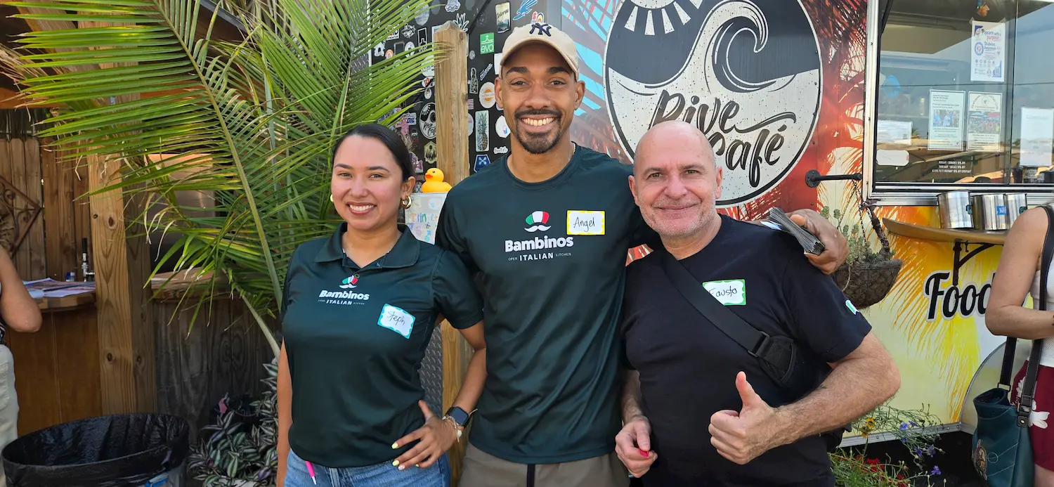 Three people stand smiling in front of a colorful food truck. Two wear green "Bambinos Italian" shirts and nametags, while the person on the right gives a thumbs-up. A palm plant is visible on the left.