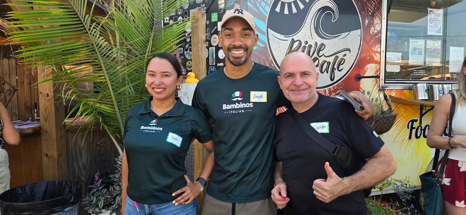 Three people stand smiling in front of a food truck. Two wear matching dark green "Bambinos Italian Inn" shirts, while the third, on the right, gives a thumbs up. The background features a surf-themed mural and a large palm leaf.