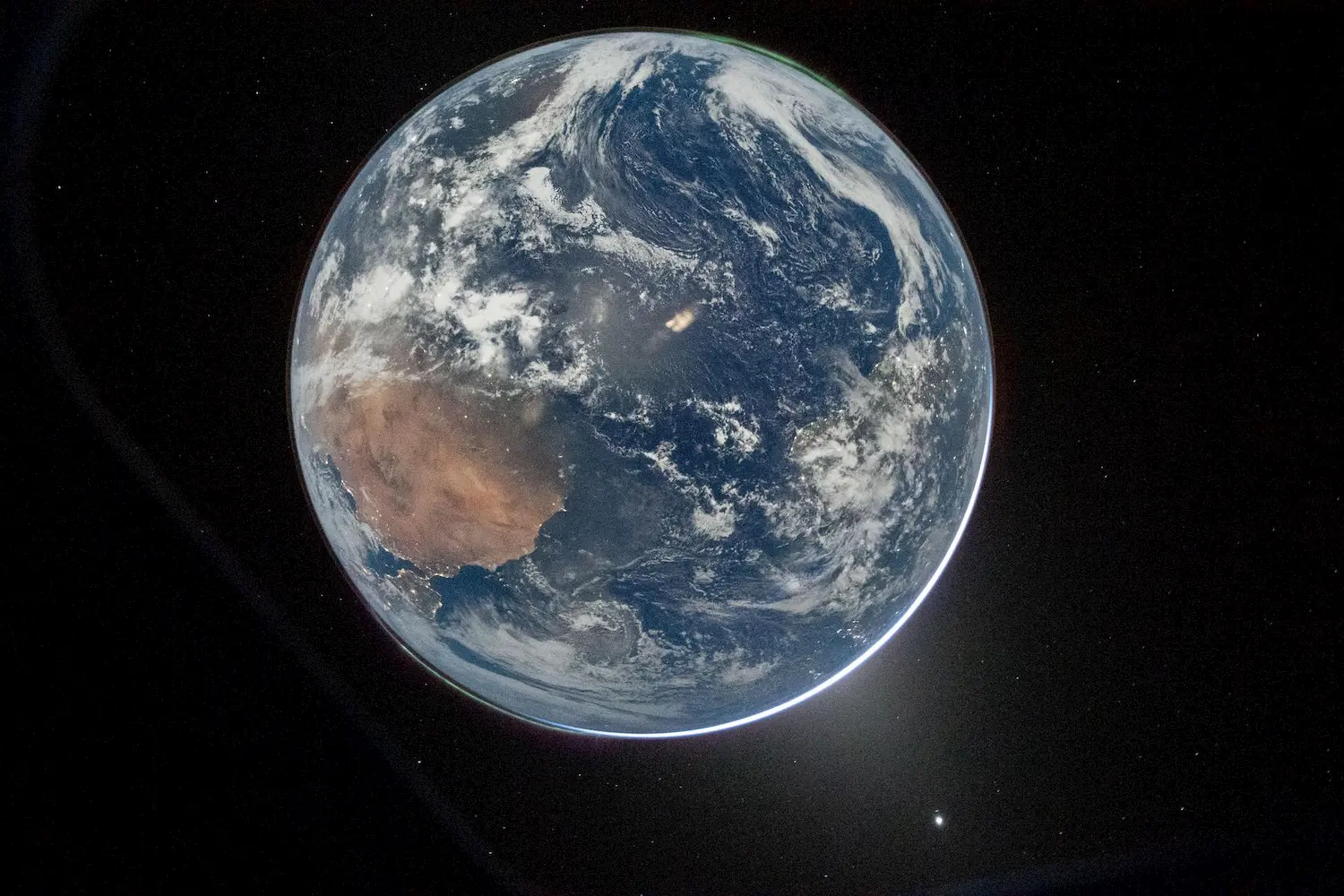 A view of planet Earth from space, showing swirling clouds, blue oceans, and the brown landmass of Africa, with the blackness of outer space and a bright spot of light visible near the bottom right.