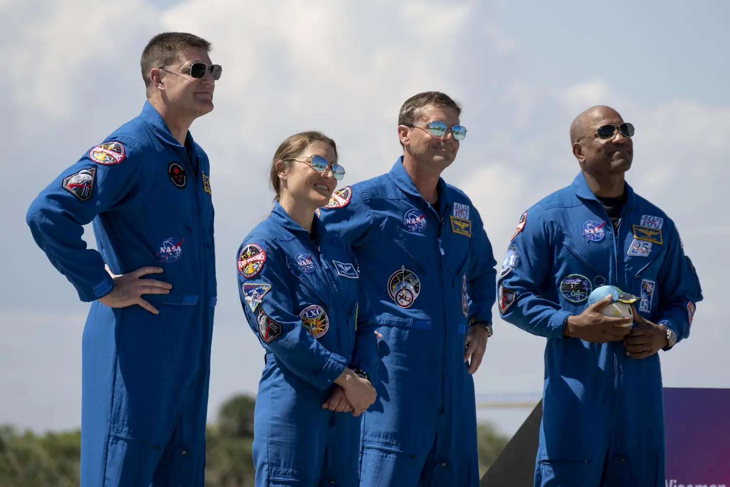 Four astronauts in blue NASA flight suits and sunglasses stand outdoors, side by side, smiling. One astronaut holds a plush toy. The sky is partly cloudy in the background.