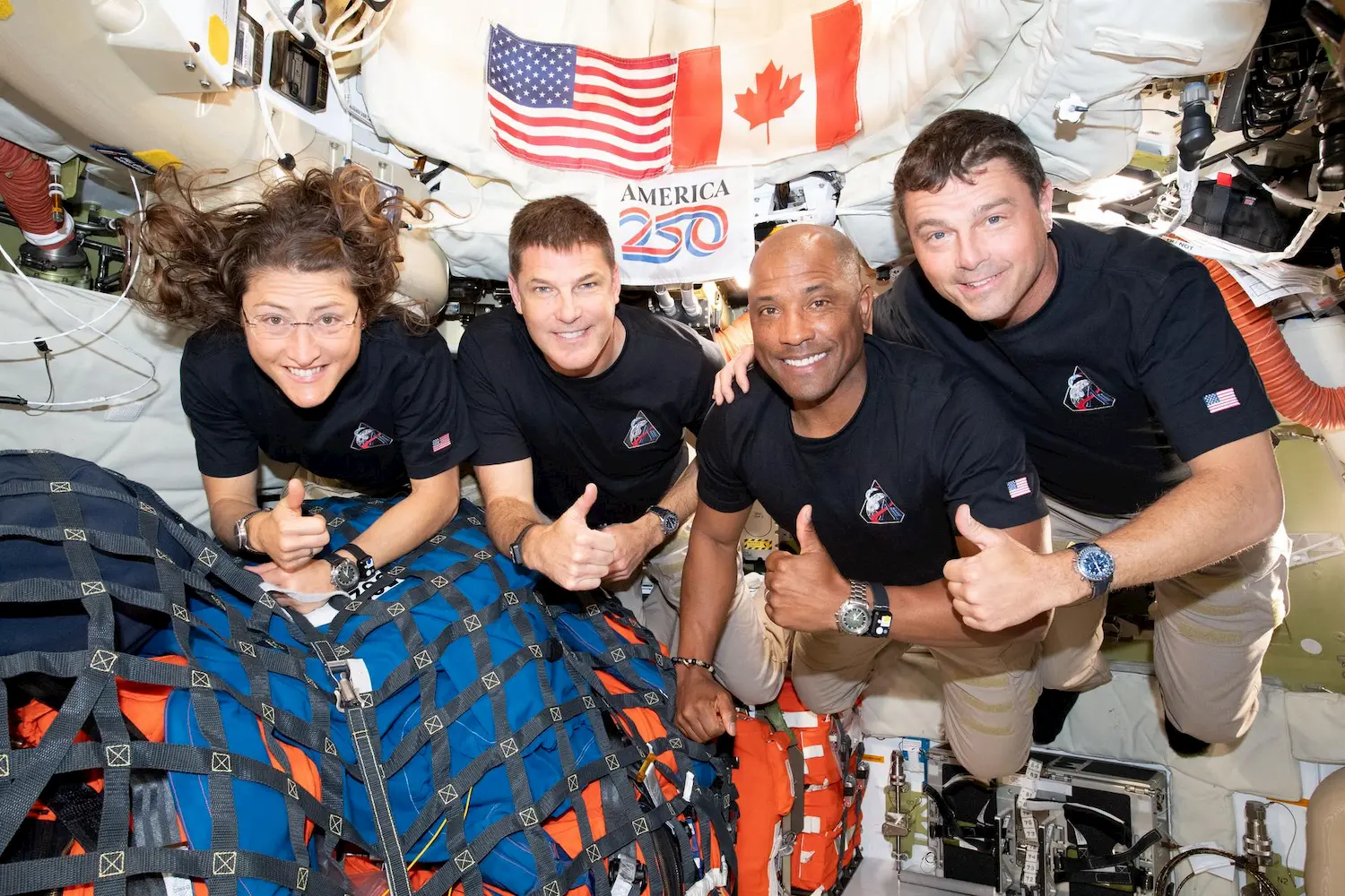 Four astronauts in black shirts smile and give thumbs up inside a spacecraft. Behind them are American and Canadian flags, equipment, and a sign reading "AMERICA 250." The environment appears bright and densely packed.