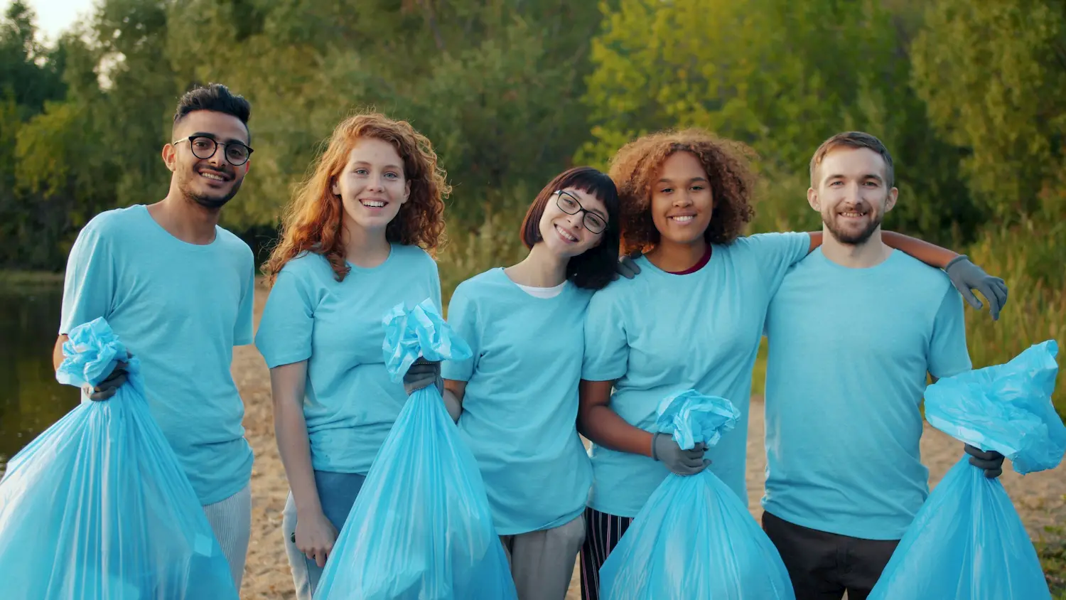A diverse group of six young adults wearing light blue shirts and holding blue garbage bags stand together outdoors, smiling, with trees and greenery in the background, participating in a cleanup activity.
