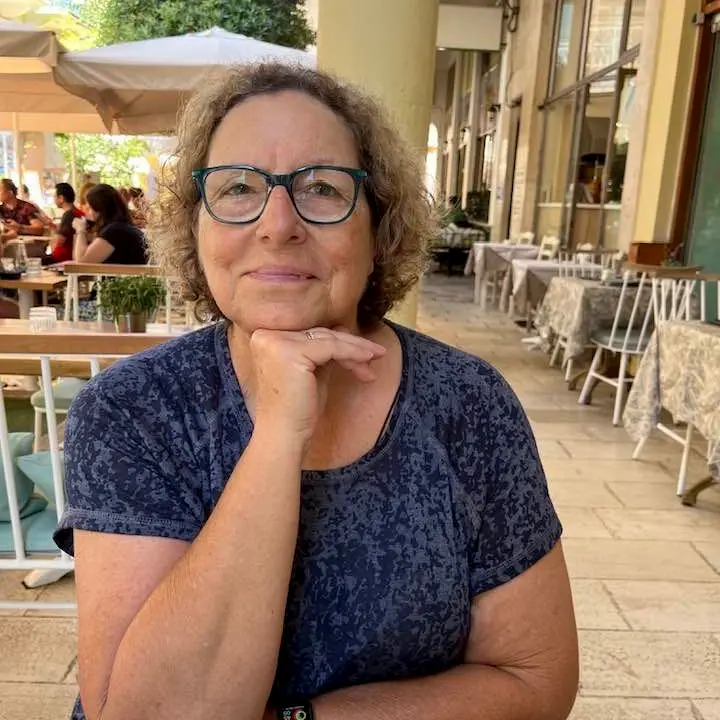 A woman with curly hair and glasses sits at an outdoor café, resting her chin on her hand and smiling slightly. She wears a blue patterned shirt, and tables and people are visible in the background.