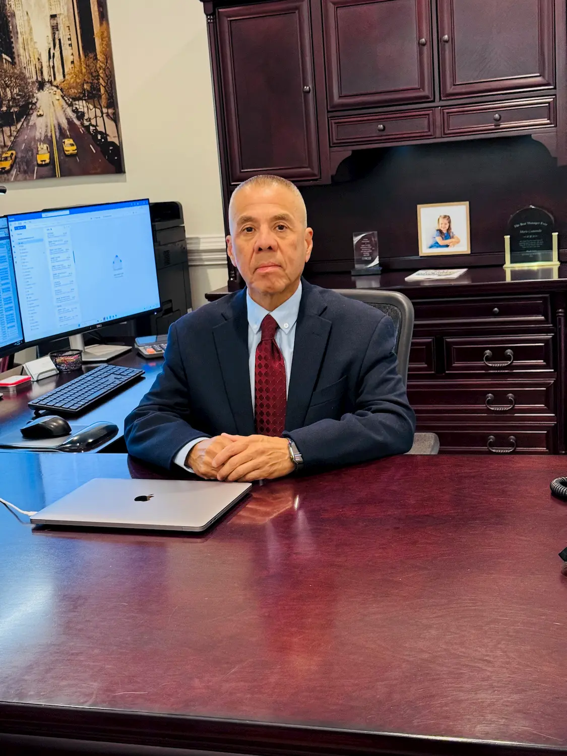 A man in a suit sits at a polished wooden desk in an office, with a laptop, computer, and framed photo behind him. He faces the camera, hands folded, with shelves and office decor in the background.