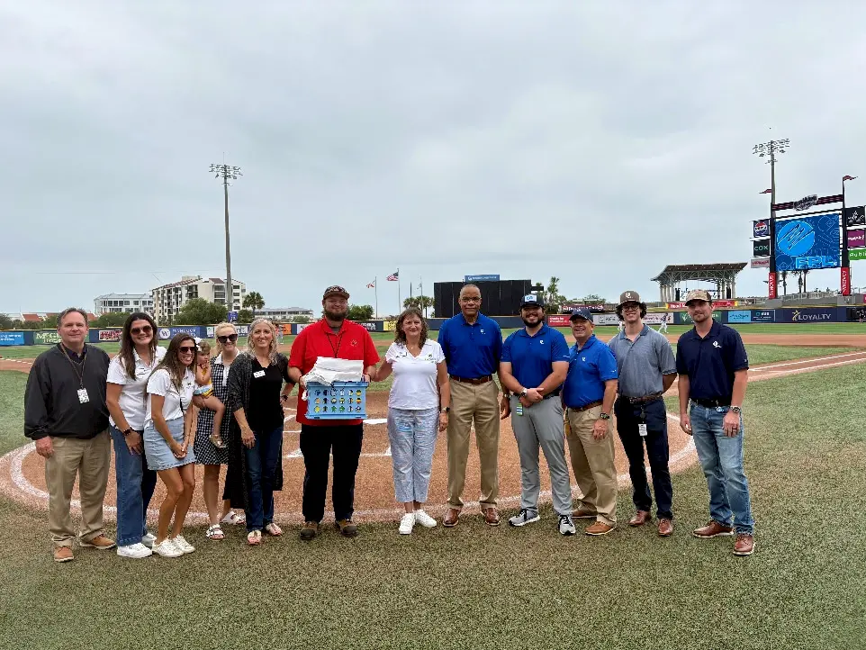 A group of twelve people stand on a baseball field near home plate, posing for a photo. Some are dressed casually, others in business attire, and one person holds a colorful display board. Stadium lights and seats are visible in the background.