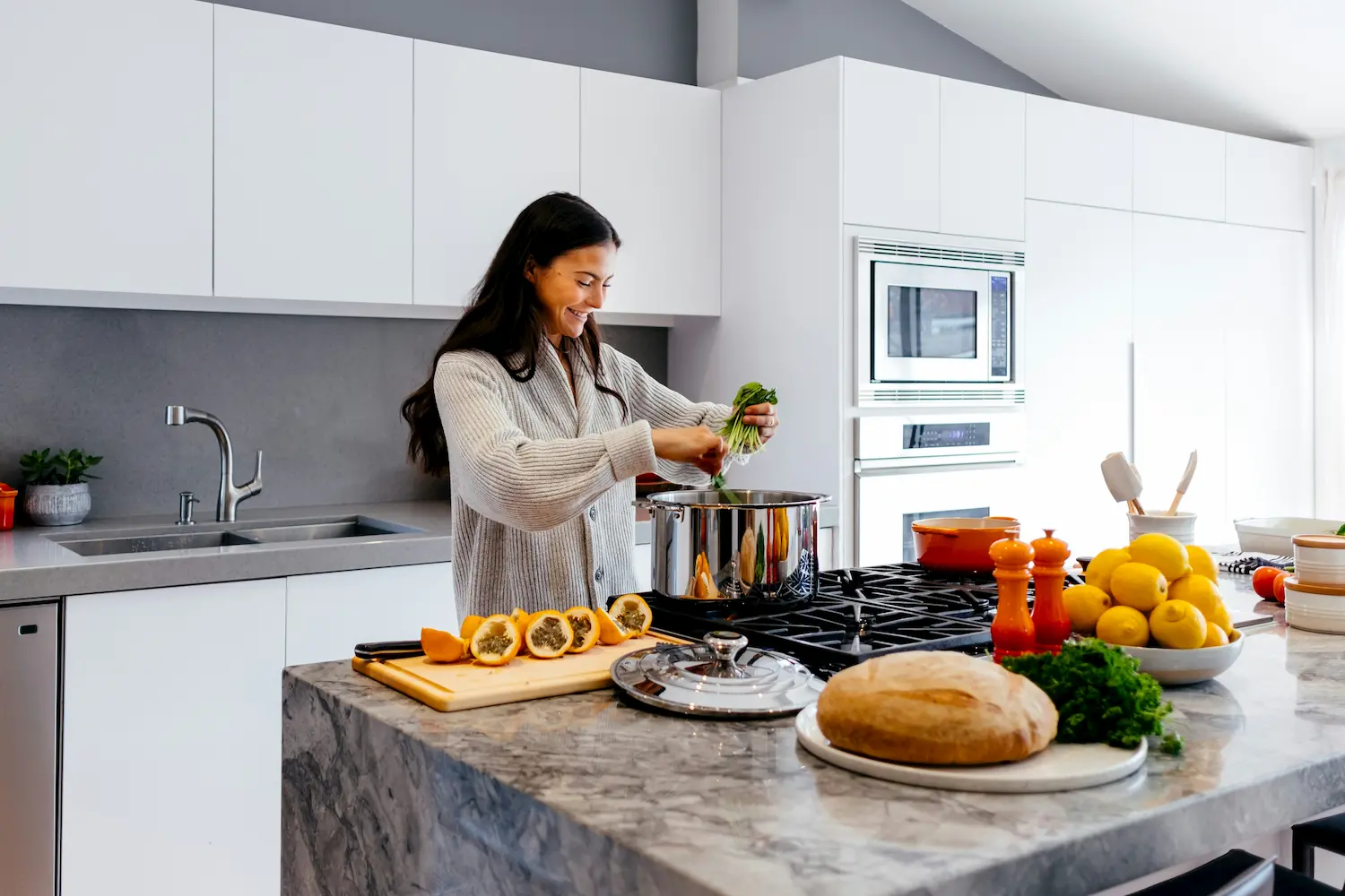 A woman smiles as she cooks in a bright, modern kitchen, adding greens to a pot on the stove. Fresh ingredients like bread, lemons, and papaya are on the marble countertop.