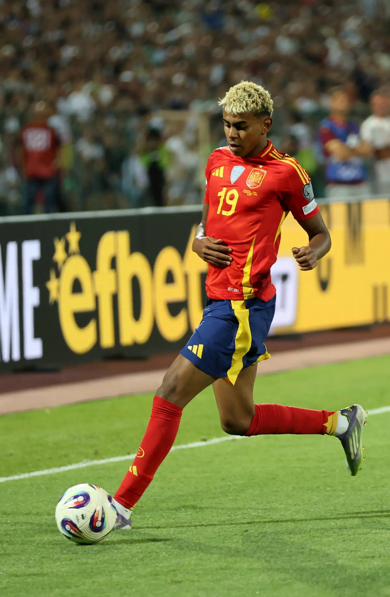 A soccer player in a red and yellow Spain jersey with the number 19 controls the ball near the sideline during a match, with a blurred crowd and advertising boards in the background.