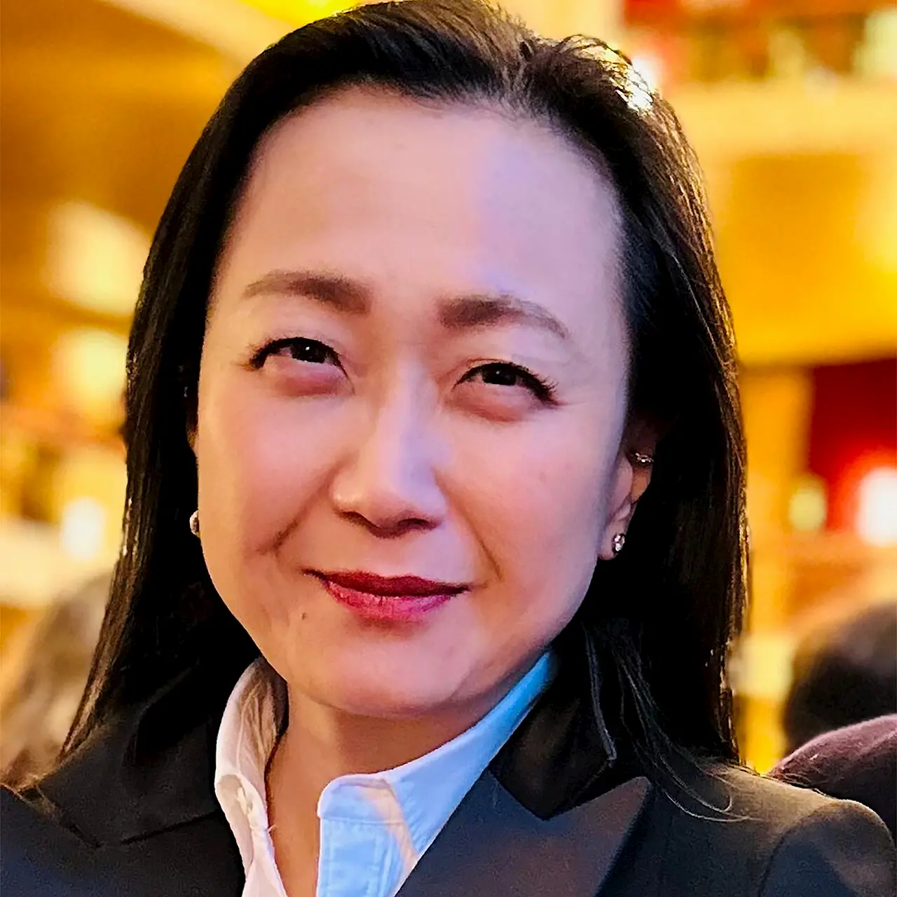 A woman with long dark hair, wearing a black blazer and white shirt, smiles softly at the camera. The background is softly blurred with warm yellow and orange tones.