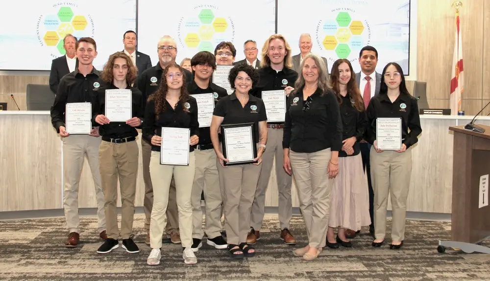 A group of students and adults pose indoors, holding certificates and smiling. They are standing in a council or boardroom, with presentation screens and a U.S. flag visible in the background.