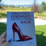 A hand with blue glitter nail polish holds the book "Two Steps Forward and Never Going Back" by Paulina Aguilar, featuring a red high heel on the cover, with a green lawn and trees in the background.