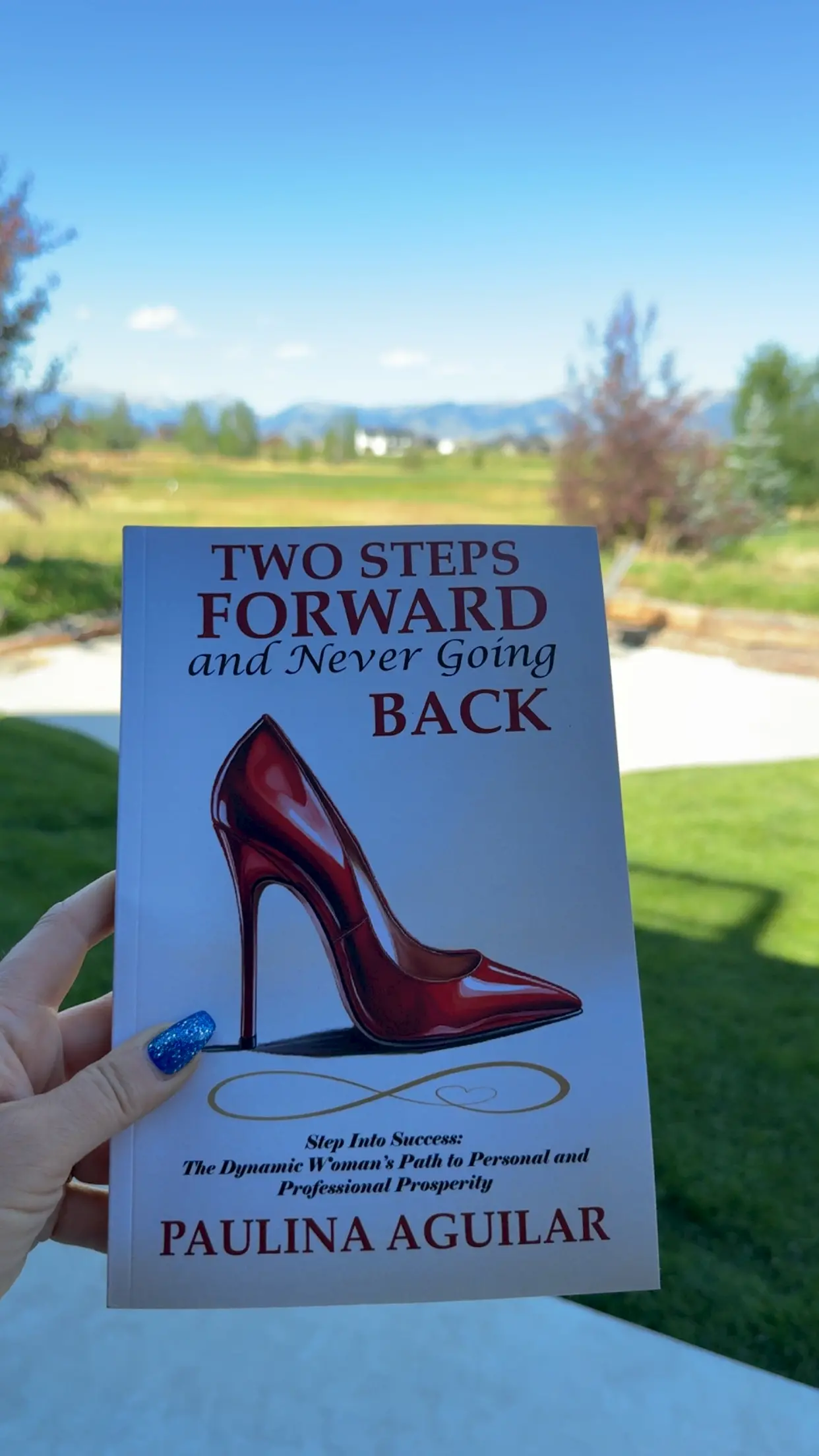 A hand with blue glitter nail polish holds the book "Two Steps Forward and Never Going Back" by Paulina Aguilar, featuring a red high heel on the cover, with a green lawn and trees in the background.