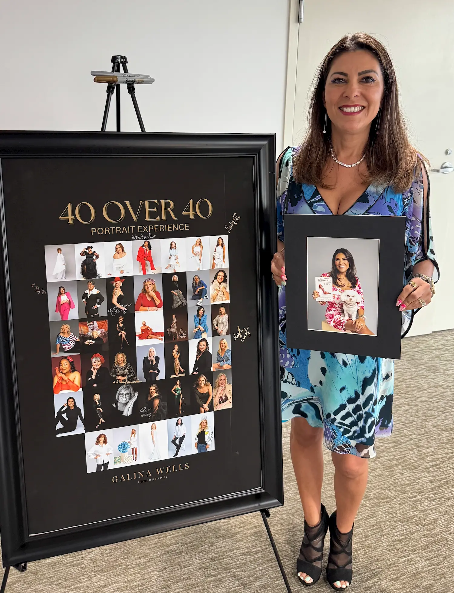 A woman in a blue dress smiles while holding a framed portrait of herself. Next to her is a large poster titled "40 Over 40 Portrait Experience" featuring photos of various women.