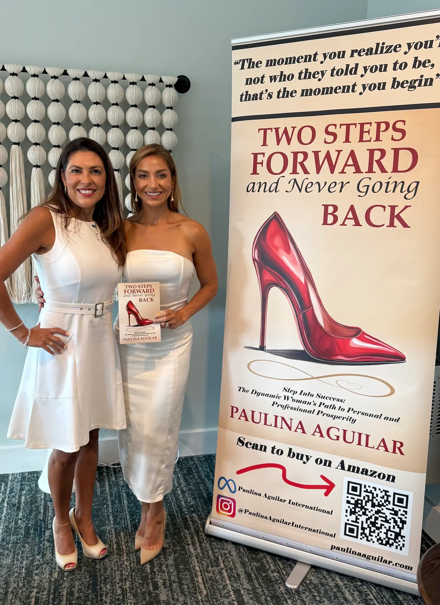 Two women in white dresses stand smiling next to a display for the book "Two Steps Forward and Never Going Back" by Paulina Aguilar. One woman holds the book; the display features a red high heel and purchasing details.
