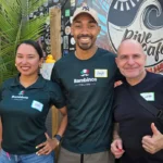 Three people stand smiling in front of the Dive Cafe food truck. The two on the left wear green Bambinos shirts, and the man on the right gives a thumbs-up. There’s a palm and colorful wall art in the background.