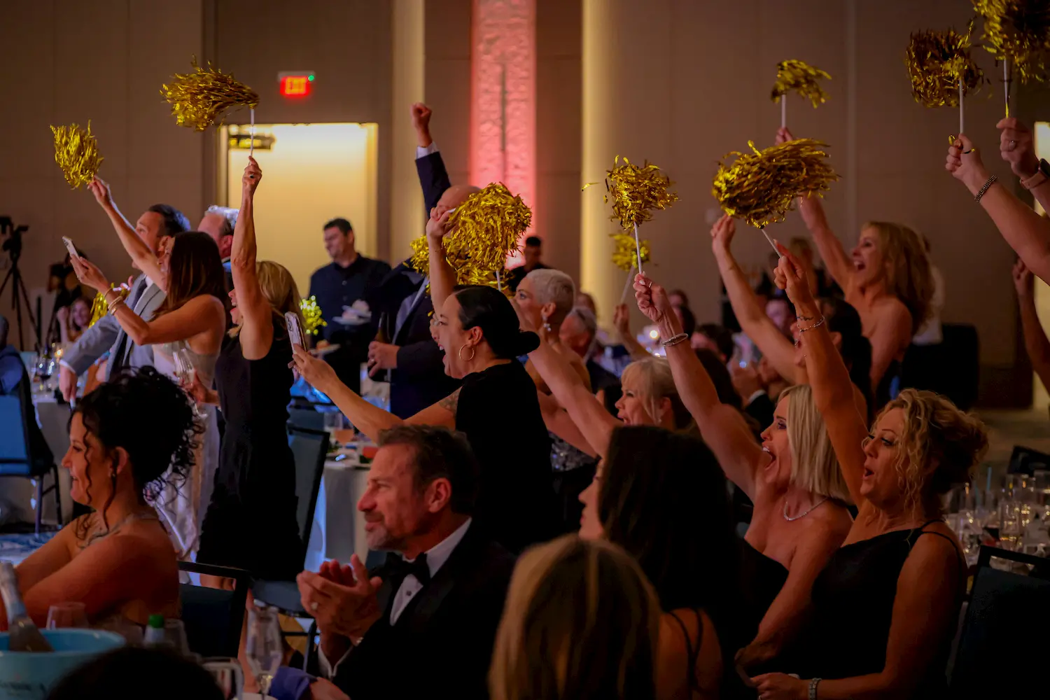 A group of people in formal attire cheer enthusiastically, waving gold pom-poms while seated at round tables during an indoor event or celebration.