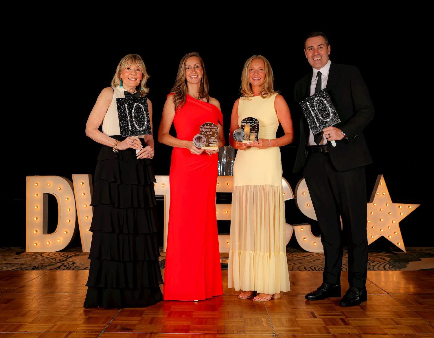 Four people dressed formally stand on a wooden floor holding awards, smiling at the camera. Large illuminated letters spelling “DWTES” with a star are displayed in the background.
