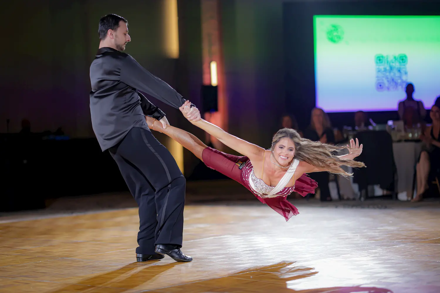A man and a woman perform an energetic ballroom dance move; the man spins the woman who is smiling and extended horizontally above the dance floor, as an audience watches in the background.