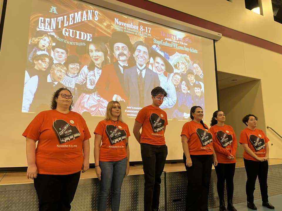 Six people wearing matching orange "Gentleman's Guide" t-shirts stand in a row on stage, with a large show poster projected behind them advertising “A Gentleman’s Guide” musical, November 8-17.