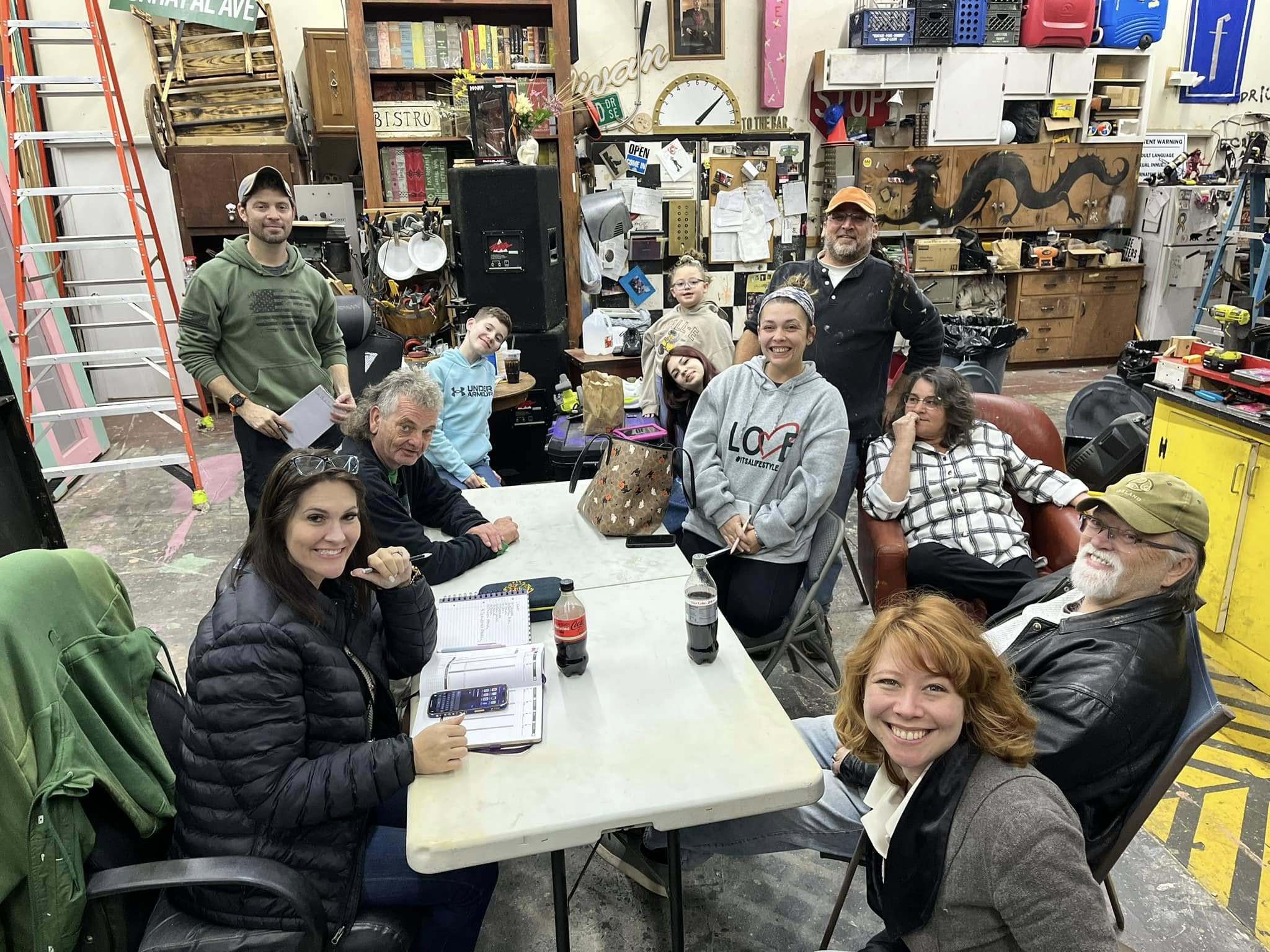 A group of people, both sitting and standing, gather around a table in a workshop or studio filled with various tools, art, and decorations. Everyone is smiling and appears to be enjoying themselves.