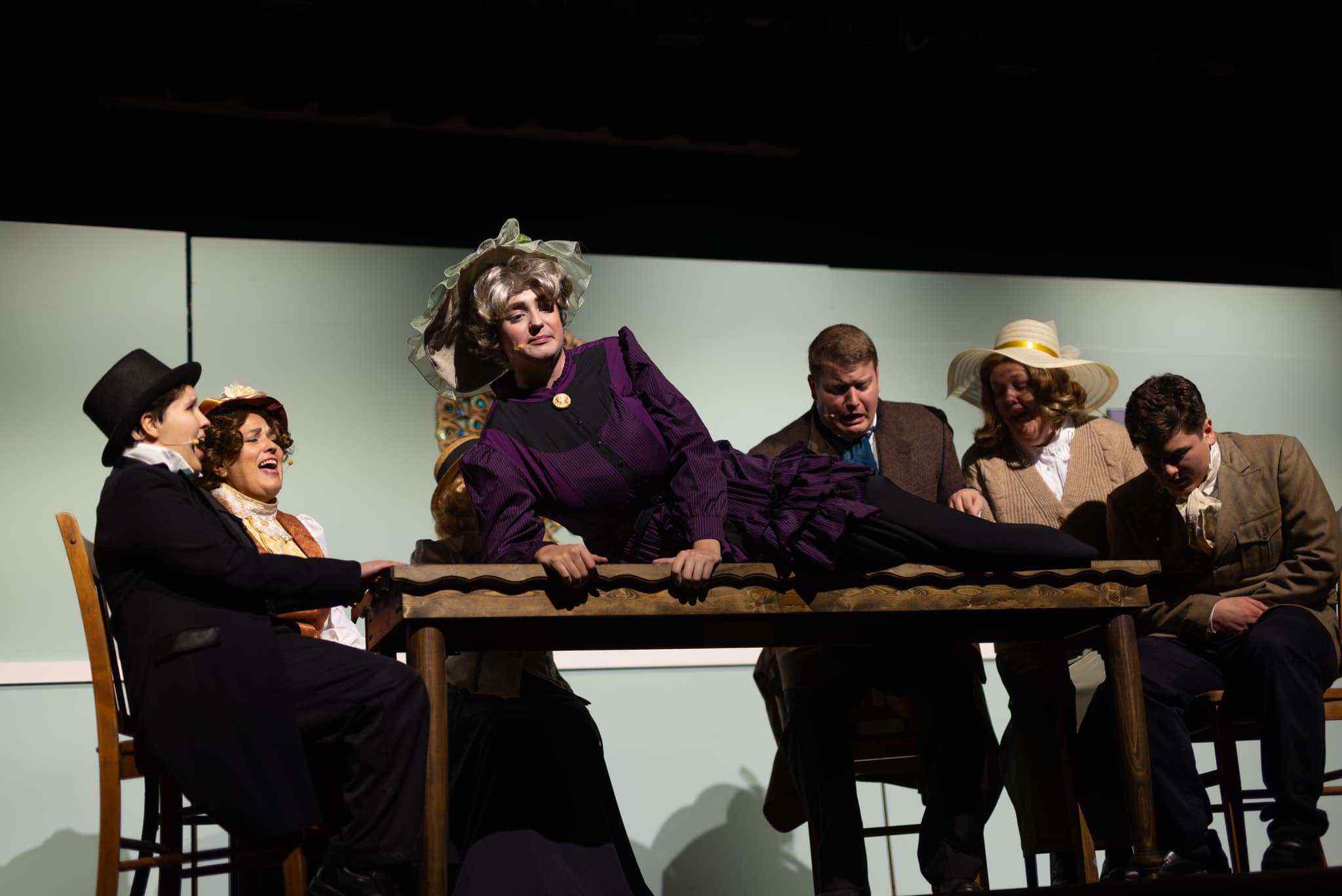 Six actors in period costumes perform a dramatic scene on stage; one person in a purple dress and large hat is lying on a table, while the others sit or kneel around, reacting emotionally.