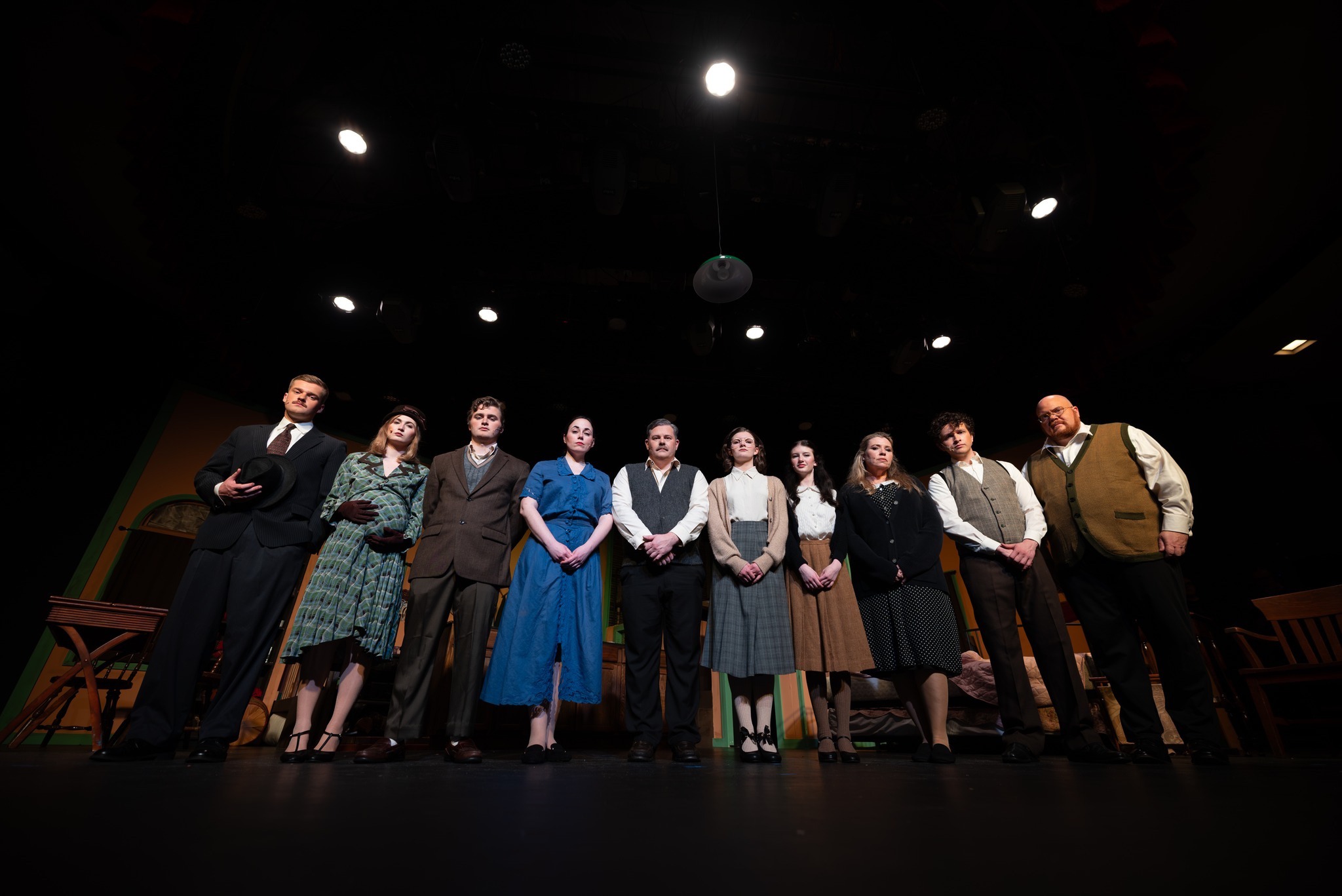 Ten people in vintage-style costumes stand in a row on a dimly lit stage, facing forward. The background includes wooden furniture and stage lights overhead.