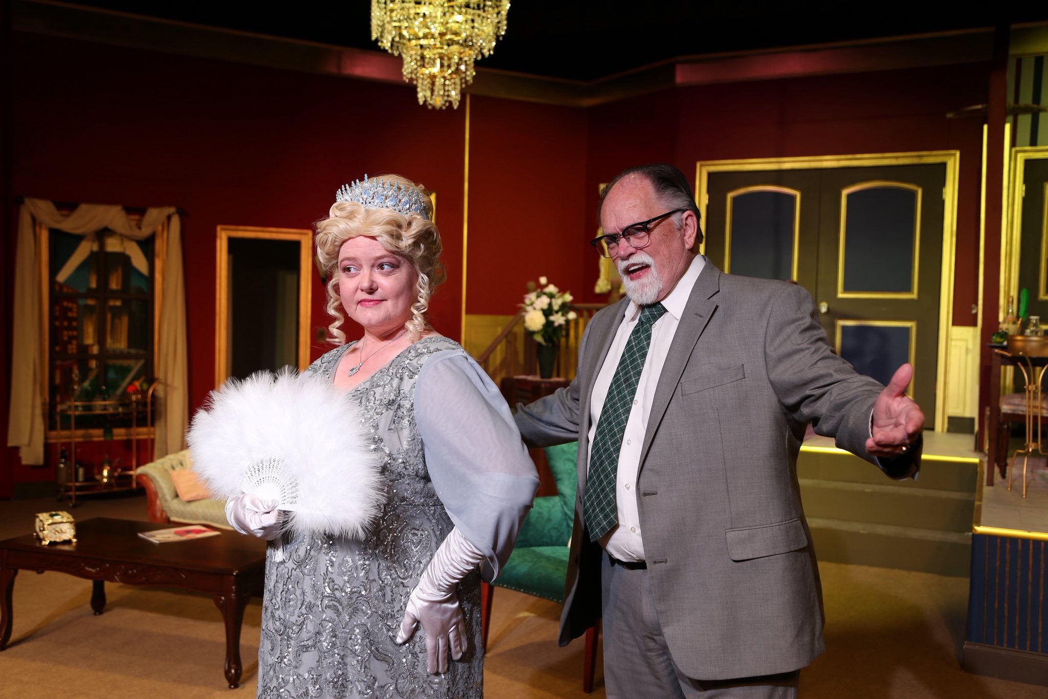 Two actors perform on stage in a lavishly decorated room. The woman in front wears a silver dress, white gloves, a tiara, and holds a white fan. The man behind her wears a gray suit and gestures with open arms.