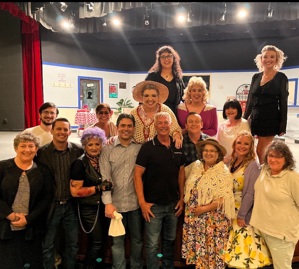 A group of fifteen adults, some in colorful costumes, pose together and smile on a theater stage with a simple set and red curtains in the background.