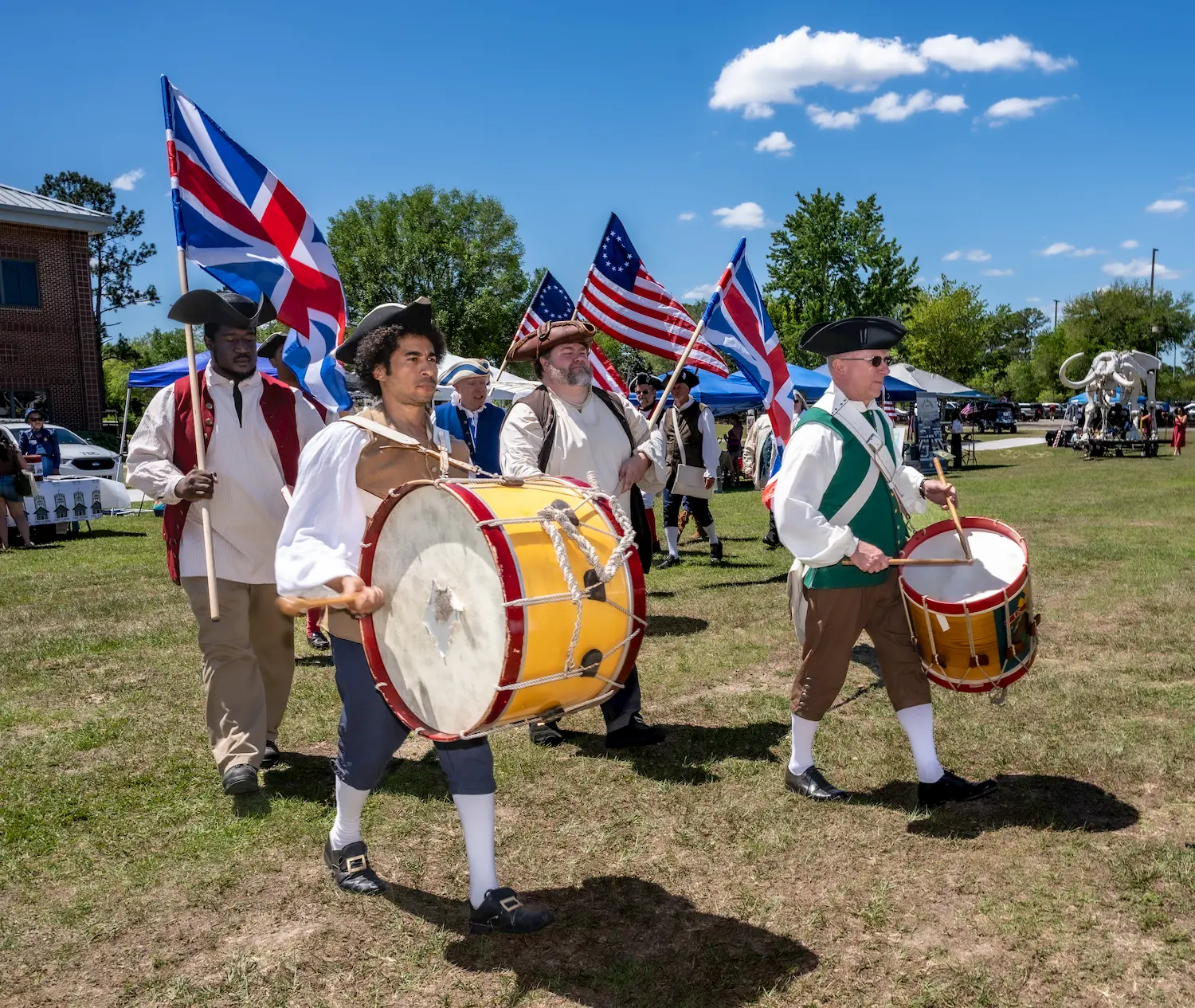 A group of people in colonial-era costumes march outdoors, playing drums and carrying British and American flags, with blue skies and trees in the background.