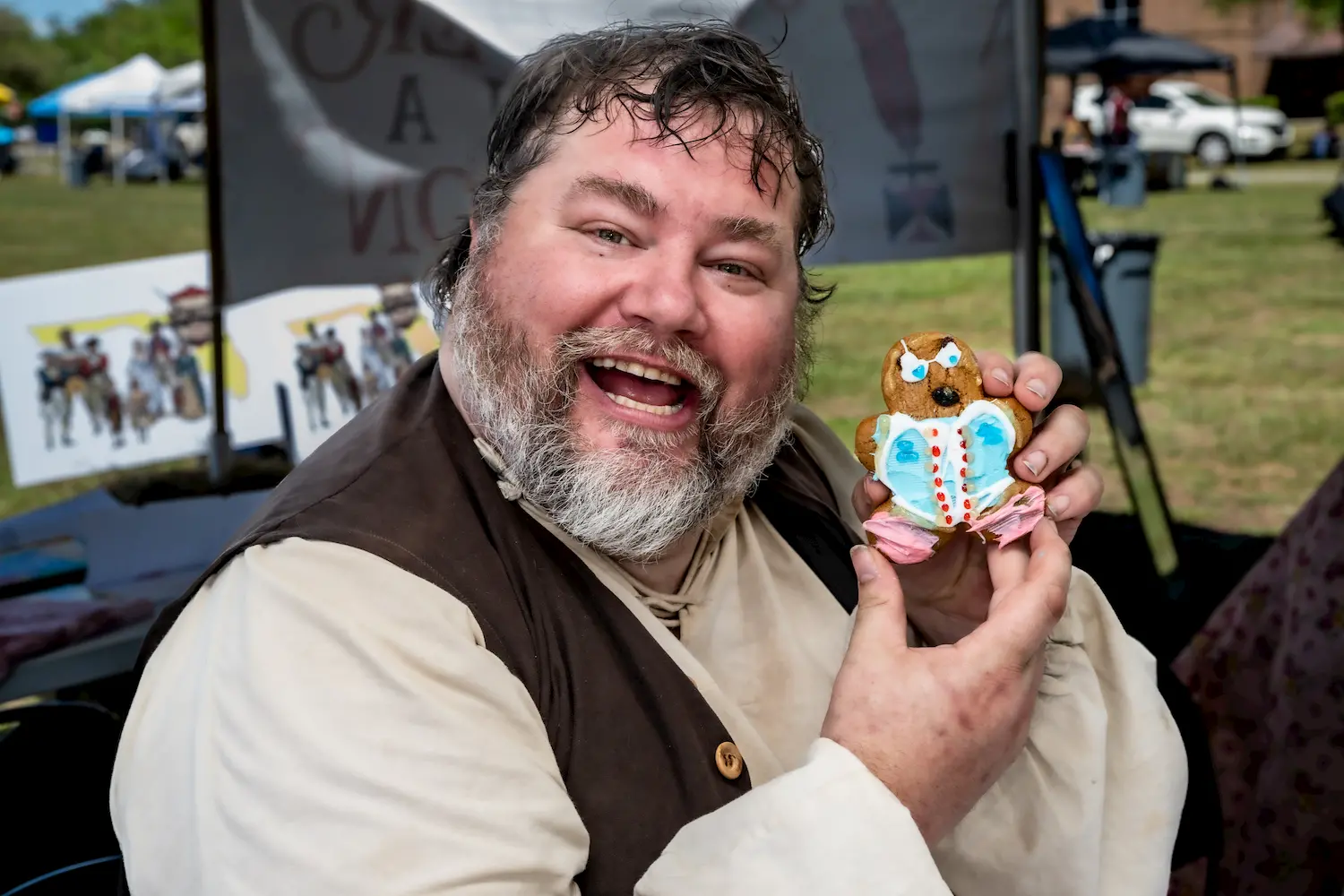 A cheerful person with a beard and medieval-style clothes smiles widely while holding a decorated gingerbread cookie shaped like a bear. Outdoor tents and people are visible in the background.