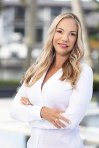 A woman with long blonde hair wearing a white blouse stands outdoors with her arms crossed, smiling confidently at the camera—embodying the power to do more through efficiency. The background is blurred with hints of buildings and trees.