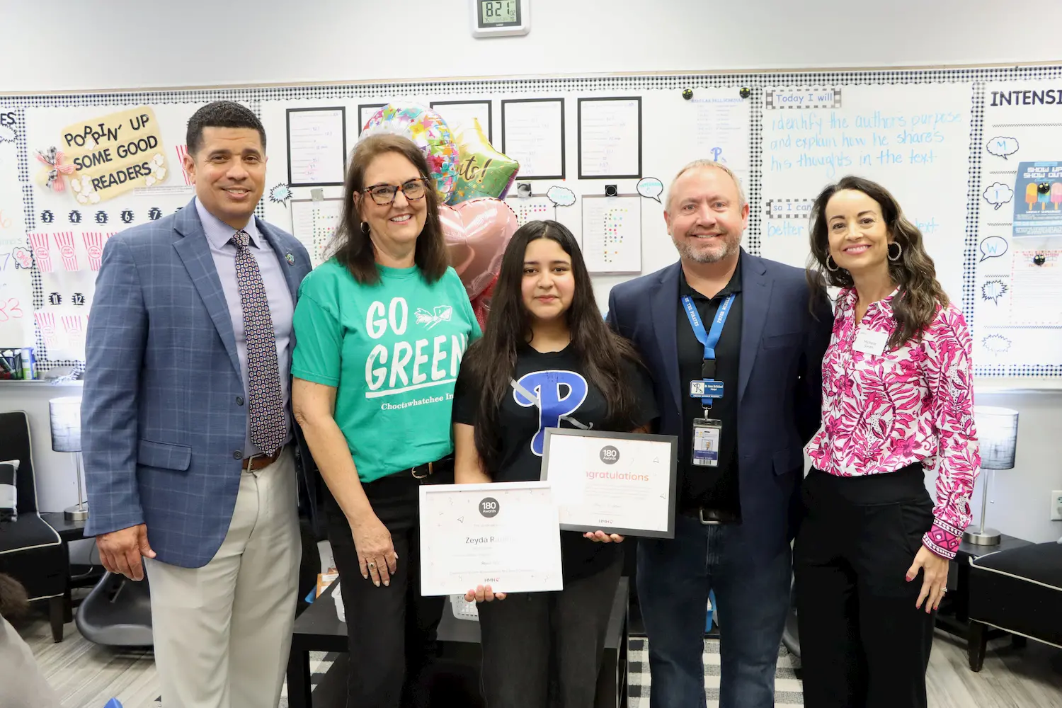 A group of five adults and one student stand together in a classroom; the student holds certificates. Balloons and classroom decorations are visible in the background, suggesting a celebration or award ceremony.