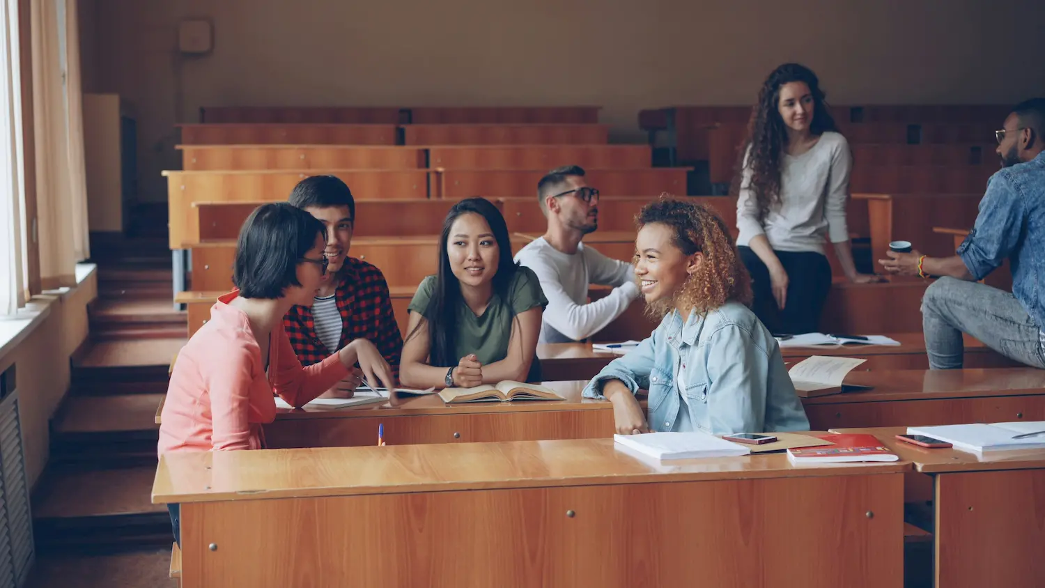 A group of college students sit and talk together in a lecture hall, with books and notebooks on their desks. Their lively discussion reflects efforts to promote peace education within positive school environments.
