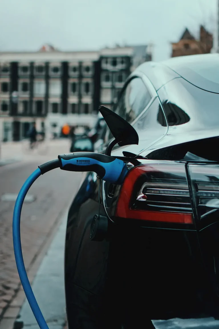 A close-up of an electric car being charged with a blue cable, parked on a city street with blurred buildings in the background—showcasing efficiency and the drive to do more with less.