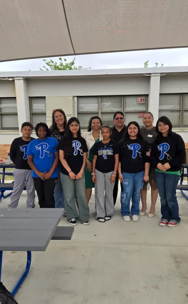 A group of ten people stand together outside a building, smiling at the camera. Most are wearing matching black shirts with a blue "P" logo. There are benches and tables in the foreground.
