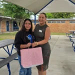 Two women stand smiling under a pavilion with blue picnic tables. One holds a pink gift bag with white dots, and the other has an arm around her. There are school buildings and greenery in the background.
