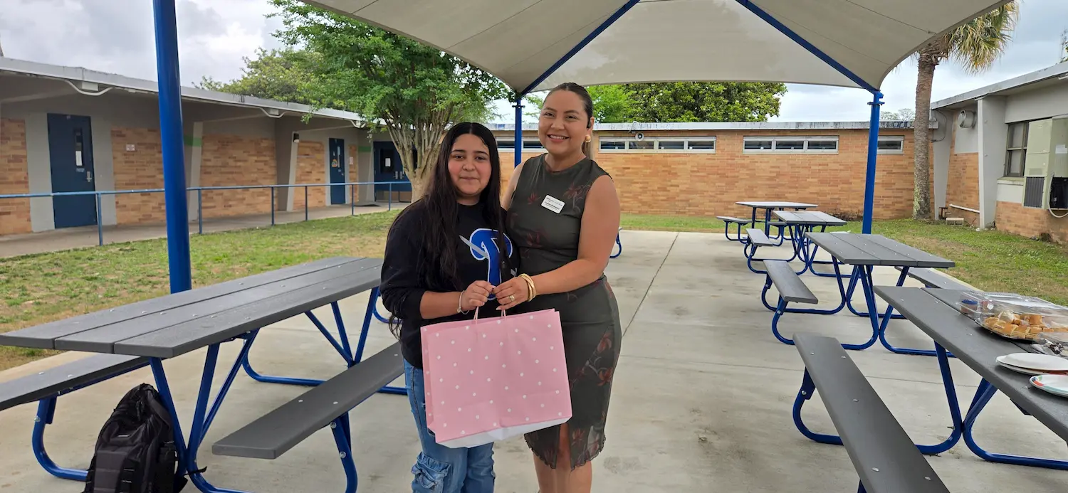 Two women stand smiling under a pavilion with blue picnic tables. One holds a pink gift bag with white dots, and the other has an arm around her. There are school buildings and greenery in the background.