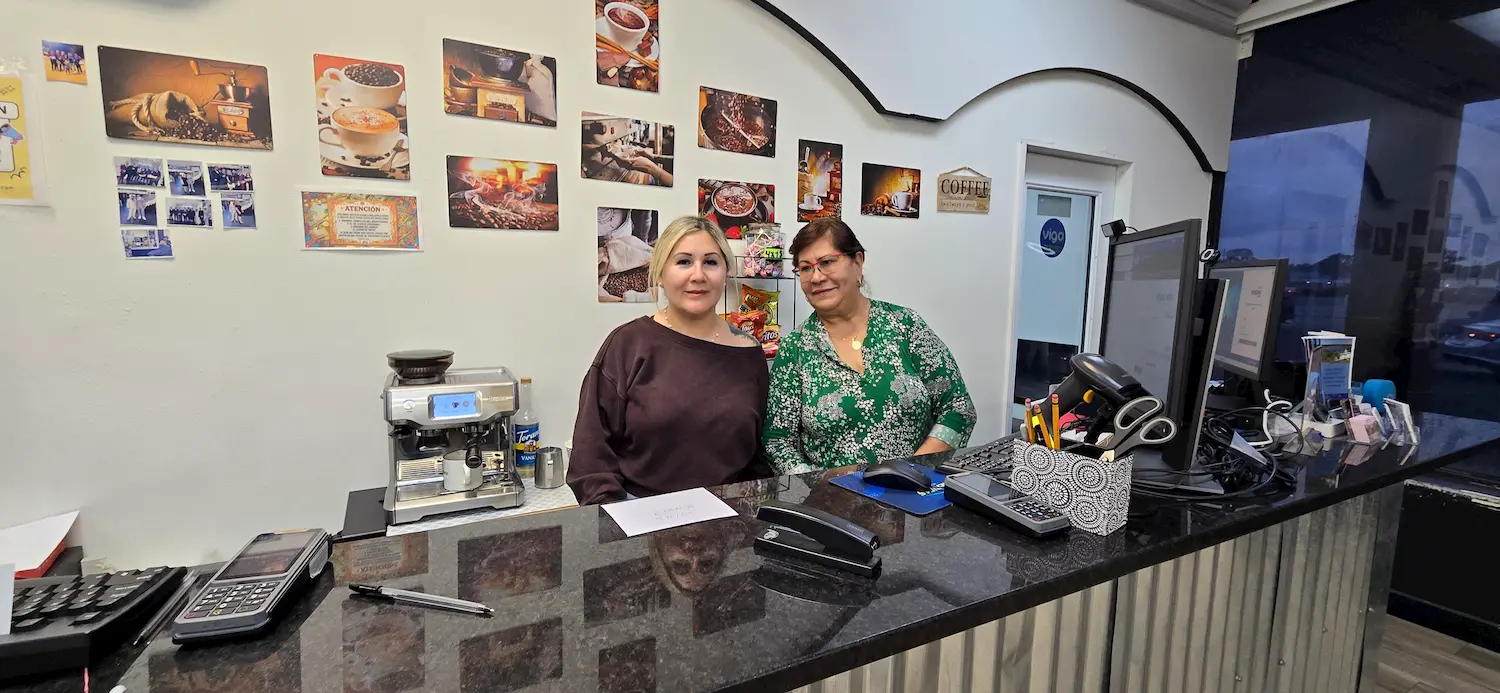 Two women stand behind salon counter with various office supplies, a cash register, and a coffee maker. The wall behind them has colorful coffee-themed photos and decorations.