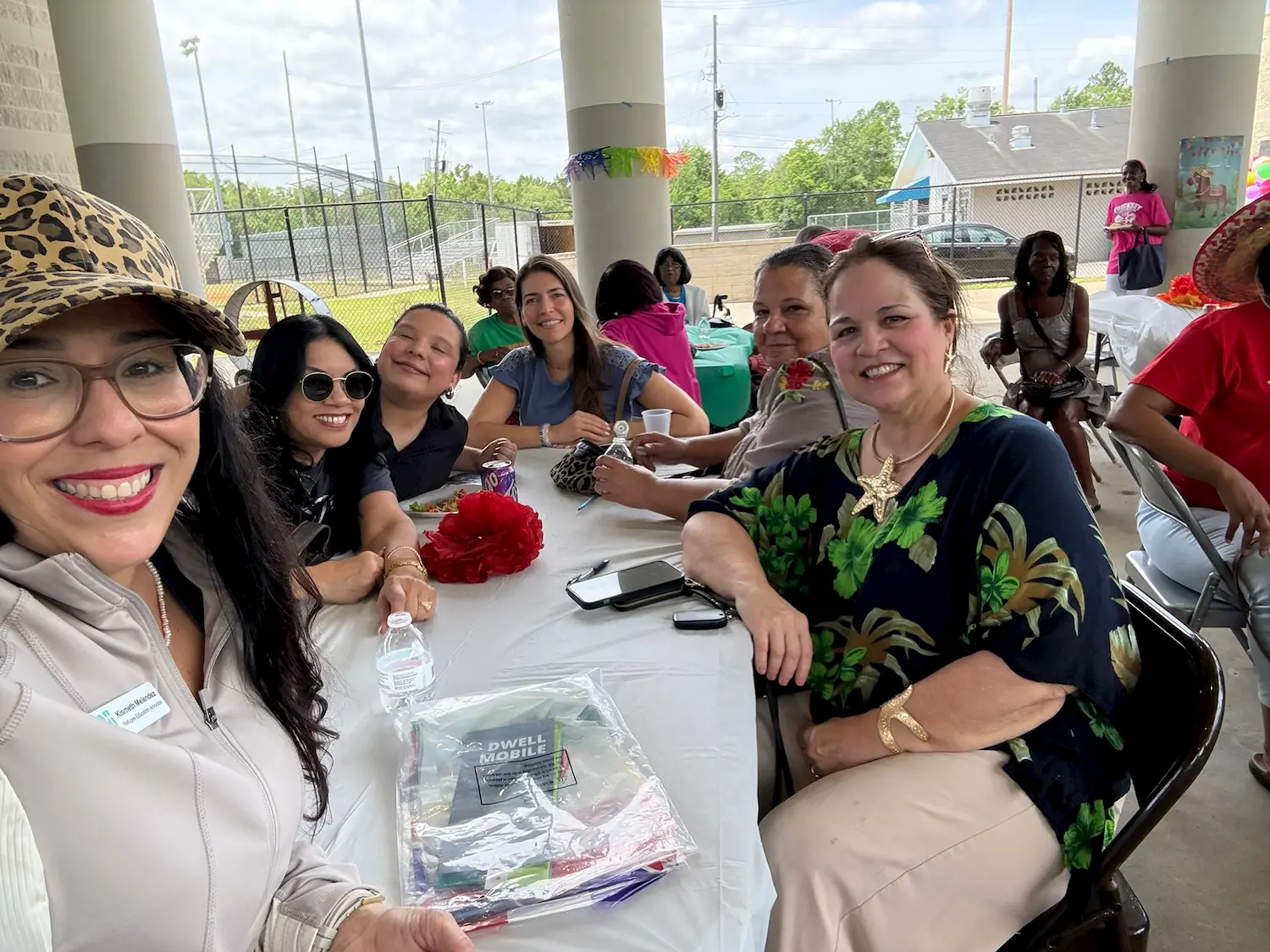 A group of smiling people sit around a table at an outdoor covered event. The table is covered with a white cloth and water bottles. There are decorations and other people in the background.